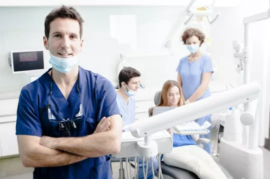 Dentist standing with his arms crossed in front of a patient sitting in a dental chair talking to two other dentists. 